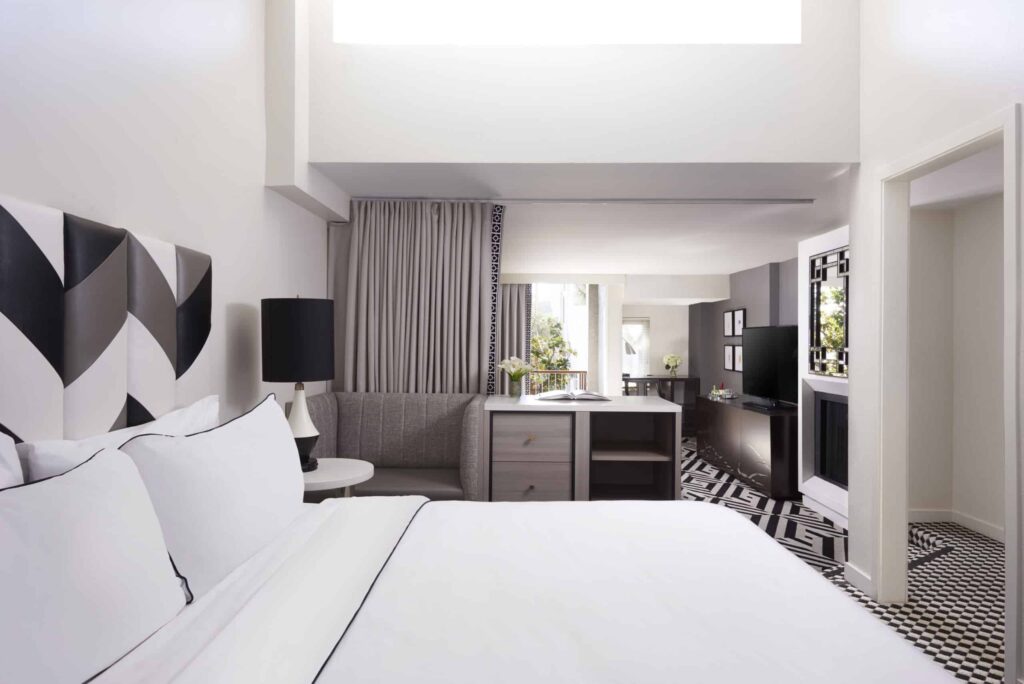 Modern guest room at the Chamberlain West Hollywood hotel with a white bed, black and white geometric decor, a sofa, desk, TV, and large window letting in natural light.