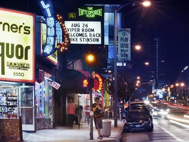 A neon-lit street at night with signs for liquor stores and The Viper Room, advertising a live performance by Mike Stinson on August 26. Cars and pedestrians are visible.