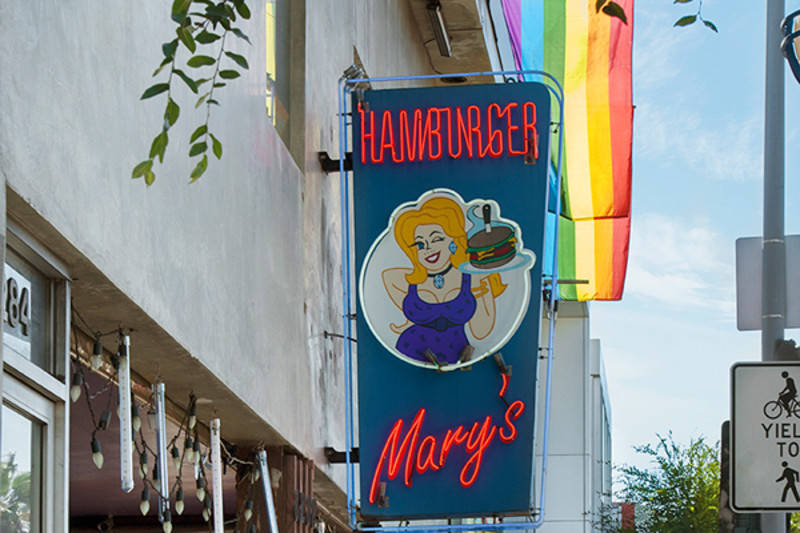 A neon sign with the words "Hamburger Mary's" and a cartoon of a drag queen holding a serving tray hangs off a building. A rainbow flag hangs behind it. Hamburger Mary's, West Hollywood, California.