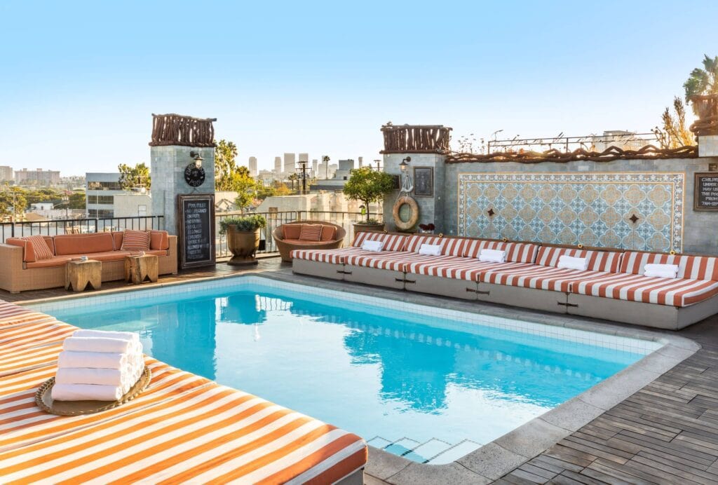 Rooftop pool area with orange and white striped lounge chairs, sofas, tile mosaic wall, and city skyline in the background under a clear sky.