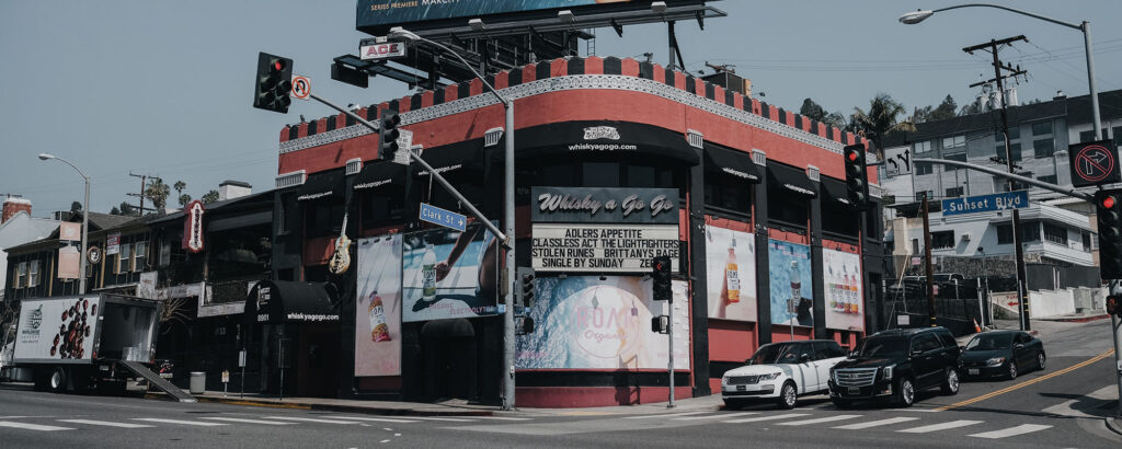 A red and black corner building with a marquee sign reading "Whisky a Go Go" on a city street, surrounded by parked cars and street signs.