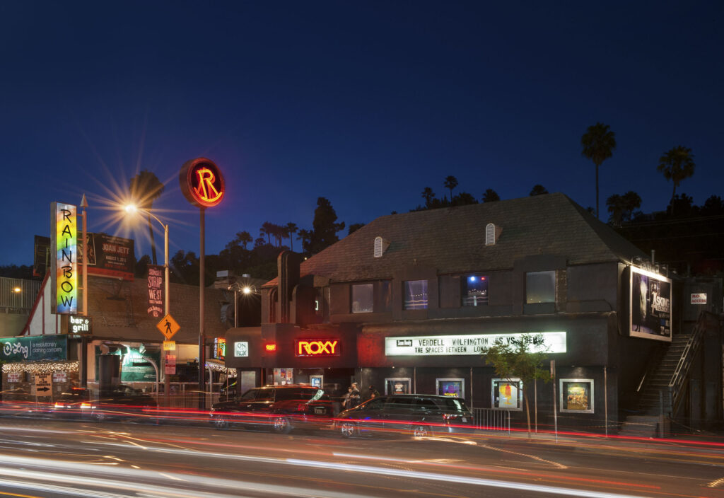 Nighttime view of The Roxy Theatre on Sunset Boulevard in Los Angeles, with neon signs lit and light trails from passing cars.