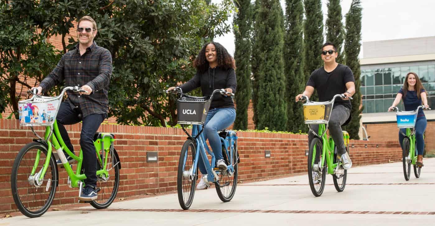 Four people ride rental bikes along a paved path bordered by red brick walls and trees on a sunny day in West Hollywood, CA
