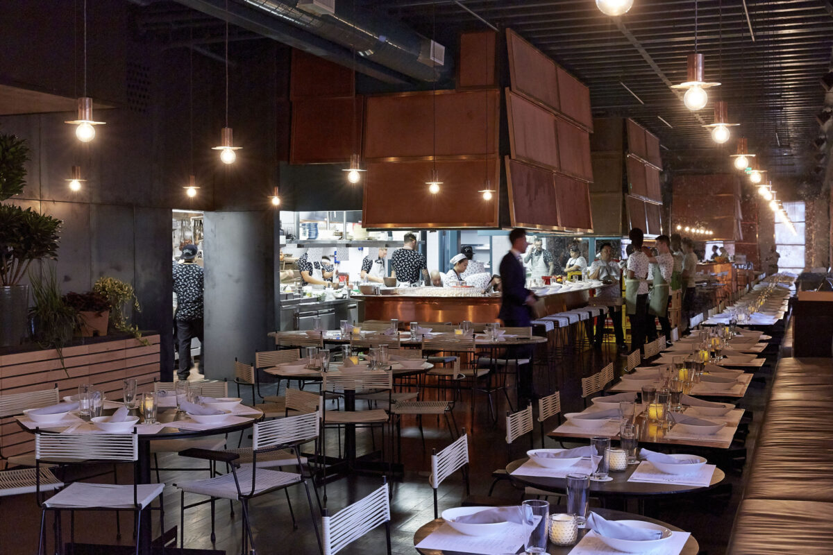 Modern restaurant interior with tables set for dining, open kitchen in the background, and staff members moving around. Warm lighting and contemporary decor are visible.