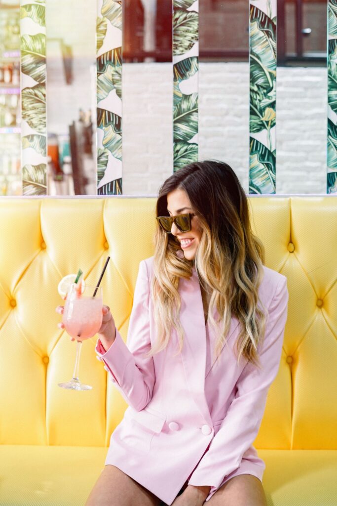 Woman in a light pink blazer sits on a yellow tufted bench, holding a pink cocktail and wearing sunglasses, with a patterned wall in the background.