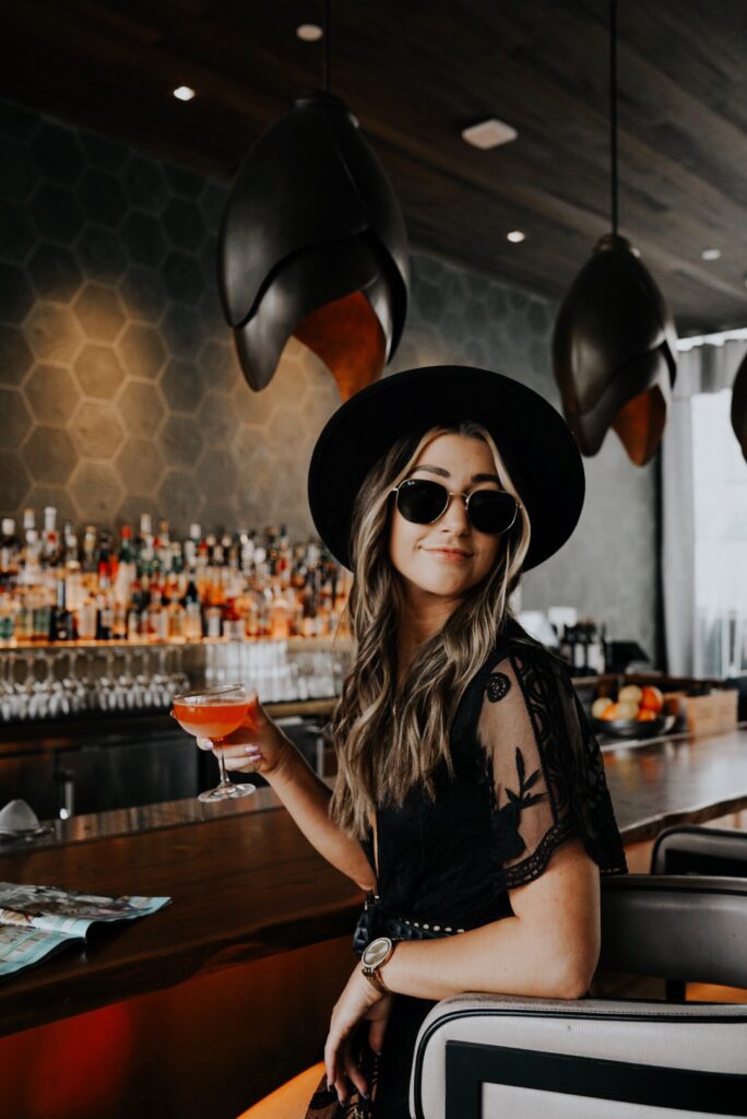 A woman wearing sunglasses and a black hat sits at a bar holding a cocktail, with shelves of liquor bottles in the background.