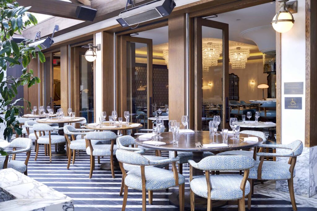 Outdoor dining area at Cecconi’s in West Hollywood, CA, with round tables and blue cushioned chairs set with glassware and napkins beside open doors leading into an elegant indoor dining room.