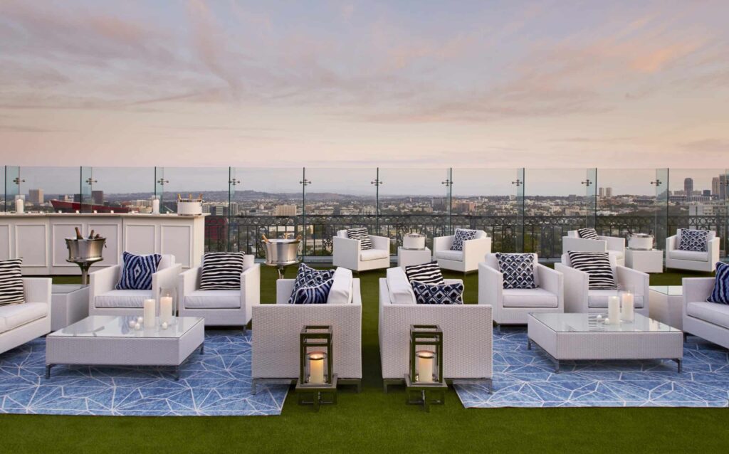 Rooftop lounge at The London West Hollywood in CA, with white sofas, glass tables, striped accent pillows and blue rugs on artificial grass, overlooking Los Angeles at sunset through a glass barrier.