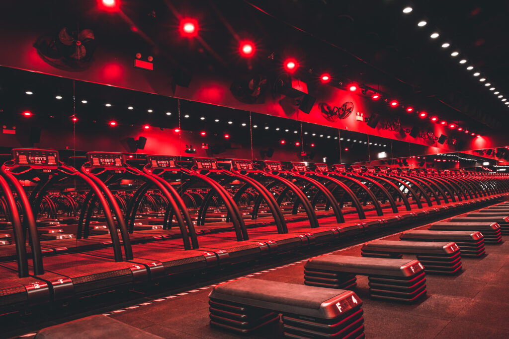 A gym studio with rows of treadmills and step platforms, illuminated by red ceiling lights and reflected in a wall of mirrors.
