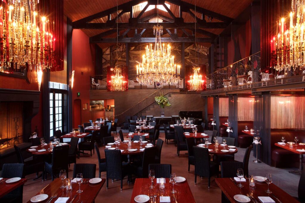 A dramatic restaurant dining room with dim lighting, dark red walls and an opulent chandelier. La Boheme, West Hollywood, California.