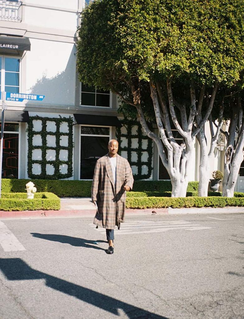 Adair Curtis, wearing a long plaid coat, gray pants and black loafers, walks across a street in front of a building with manicured hedges and large trimmed trees.
