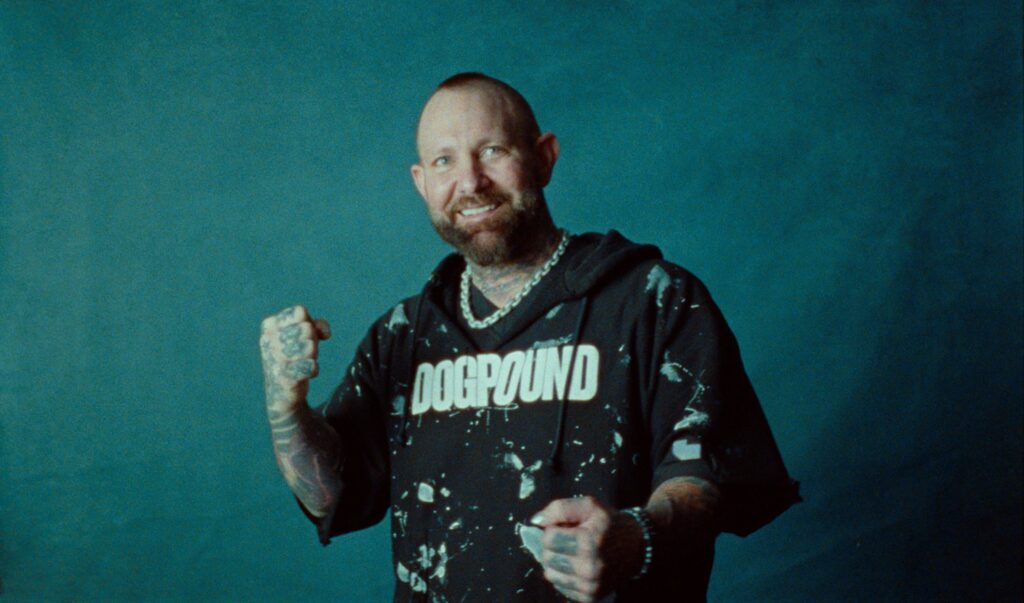 A tattooed man with a beard and shaved head poses with clenched fists, wearing a "DOGPOUND" shirt and chain necklace against a blue background.