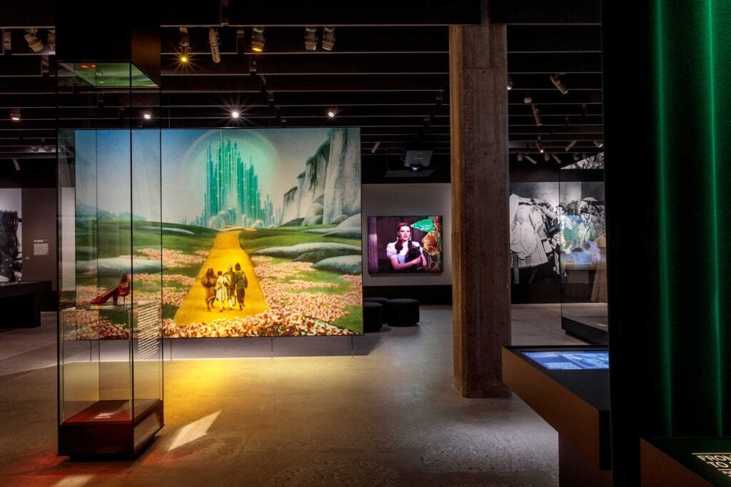 A museum exhibit features a large display of the Emerald City scene from "The Wizard of Oz," with related photos and artifacts in a modern, dimly lit space.