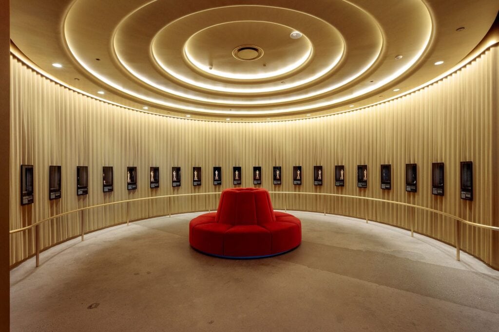 Circular room with concentric ceiling lights, lined with wall-mounted display screens, and a central red round seating area.