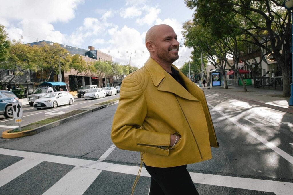 A person wearing a yellow jacket crosses a city street at a crosswalk, with cars and trees lining the road in the background.