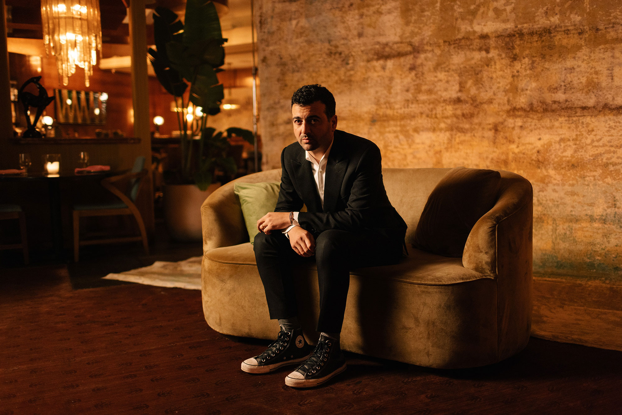 A man in a black suit and Converse sneakers sits on a tan velvet sofa in a warmly lit room with rustic walls and ambient lighting.