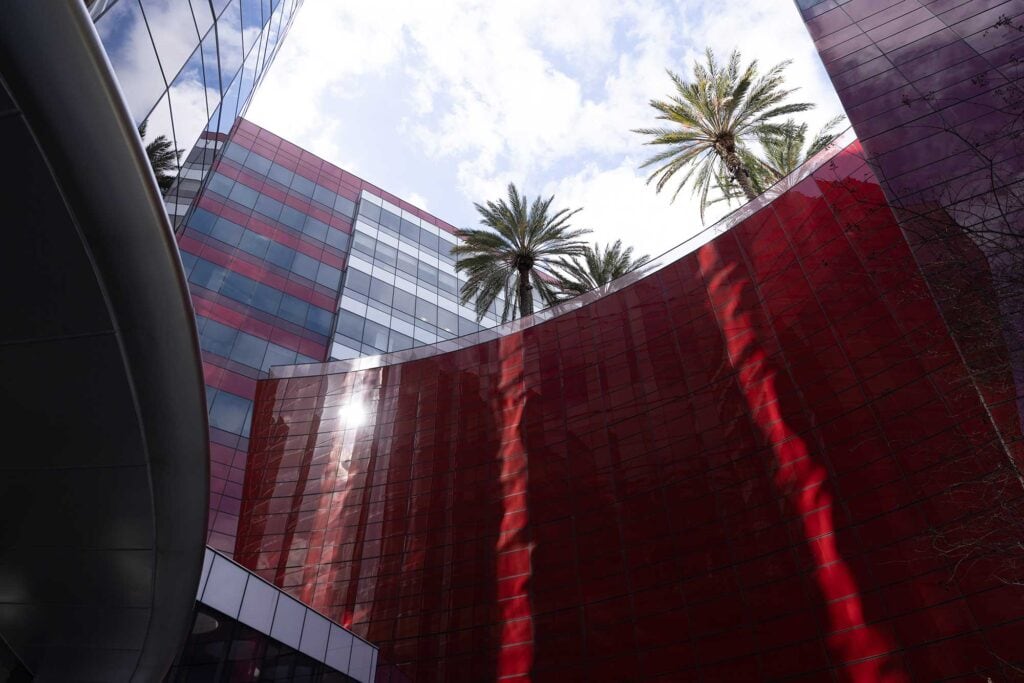 Curved red-tiled building facade with mirrored windows, adjacent structures, and palm trees rising above against a partly cloudy sky.