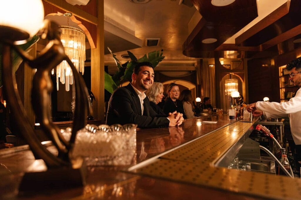 Three people sit at a bar counter smiling, while a bartender prepares drinks in a warmly lit, elegant setting with chandeliers and decorative elements.