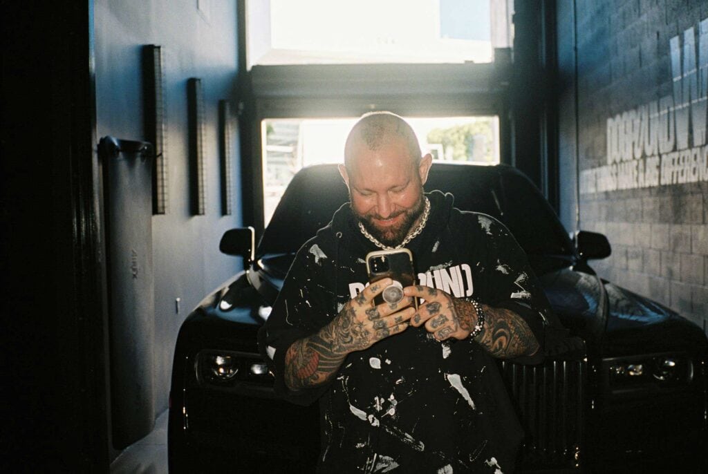 A tattooed man stands in front of a black car inside a garage, smiling as he looks at his phone. Sunlight shines through the open entrance behind him.