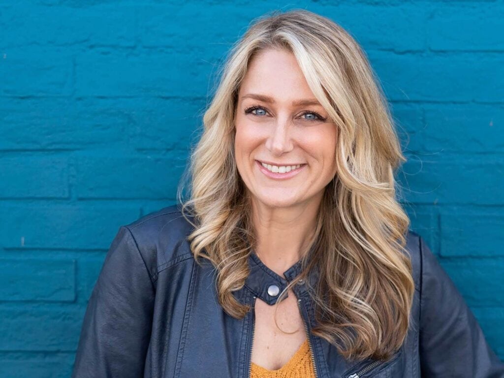 A woman with long blonde hair smiles at the camera, wearing a black leather jacket and standing in front of a blue brick wall.