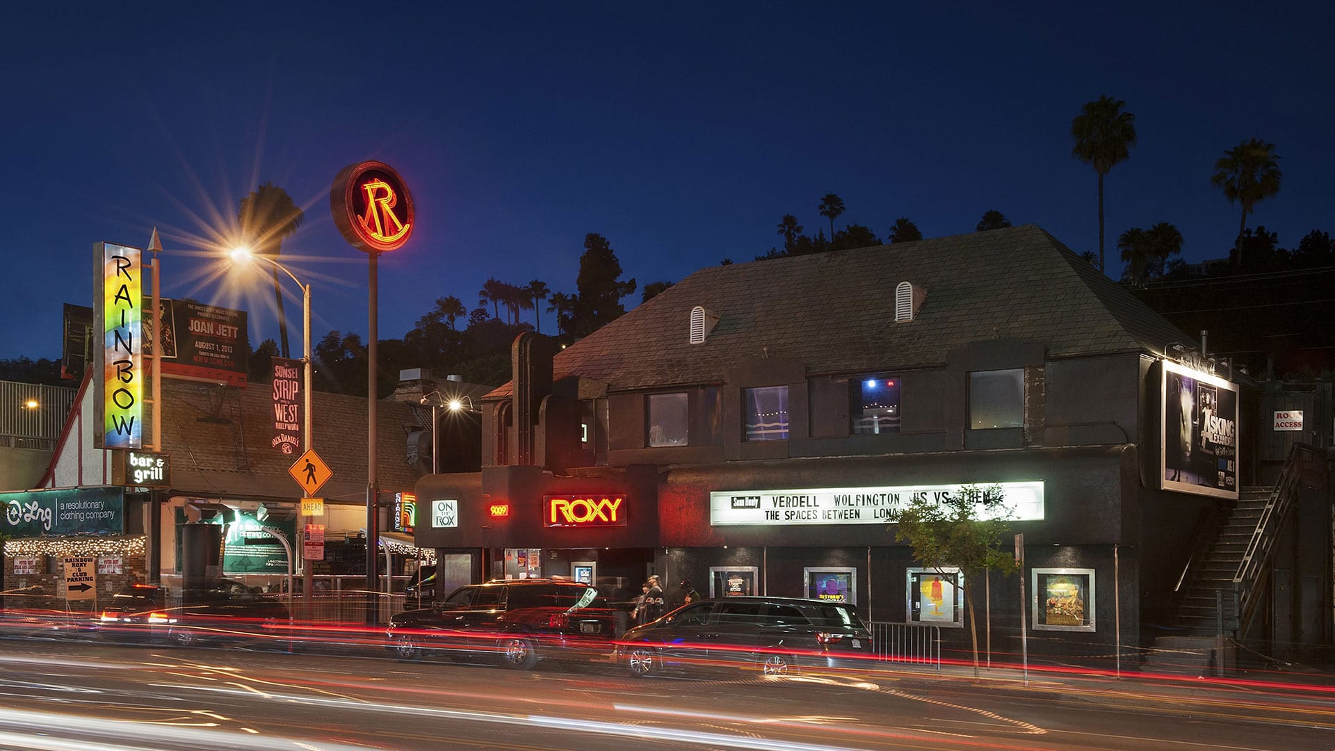 Night view of The Roxy Theatre in Los Angeles with illuminated signs, parked cars, and light trails from traffic on Sunset Boulevard.