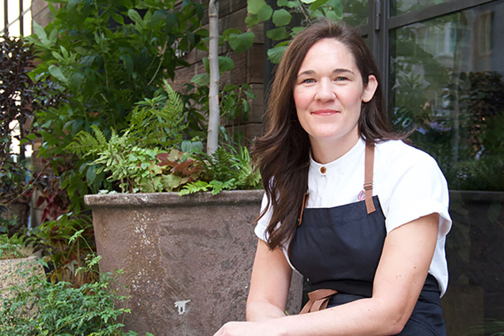 A woman with brown hair, Chef Ginger Pierce, sits in a garden environment wearing a black chef's apron.