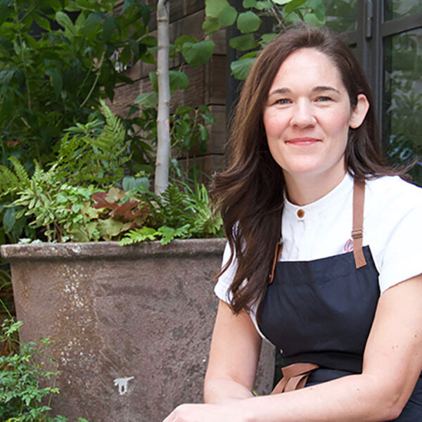 A woman with brown hair, Chef Ginger Pierce, sits in a garden environment wearing a black chef's apron.