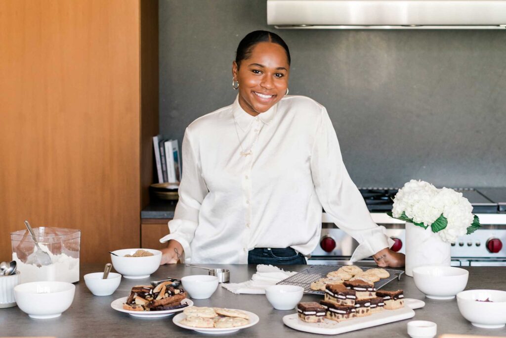 A woman wearing a white button-down shirt stands in a modern kitchen behind a counter filled with desserts. West Hollywood, California.