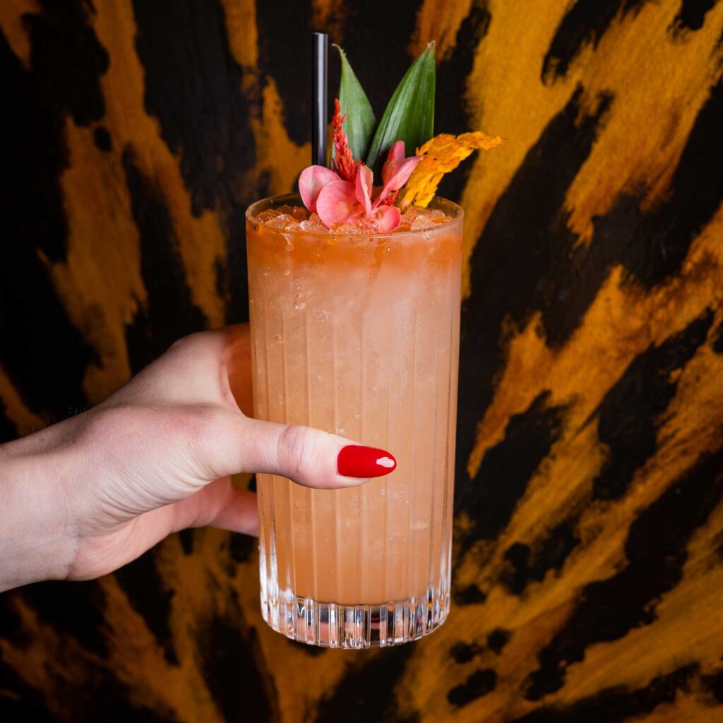 A woman's hand with red fingernails holds a cocktail in a highball glass in front of an animal-print wall.
