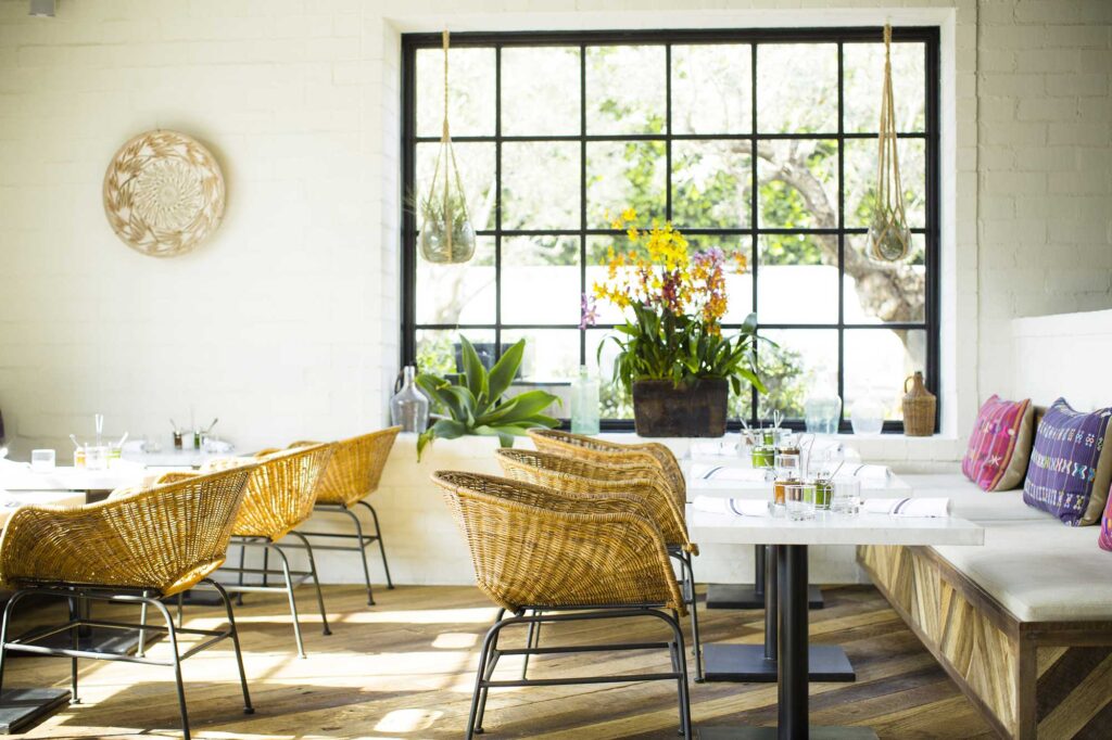 Natural light floods into an airy, white restaurant dining room featuring large windows, basket-style seats and dangling plants. Gracias Madre, West Hollywood, California.