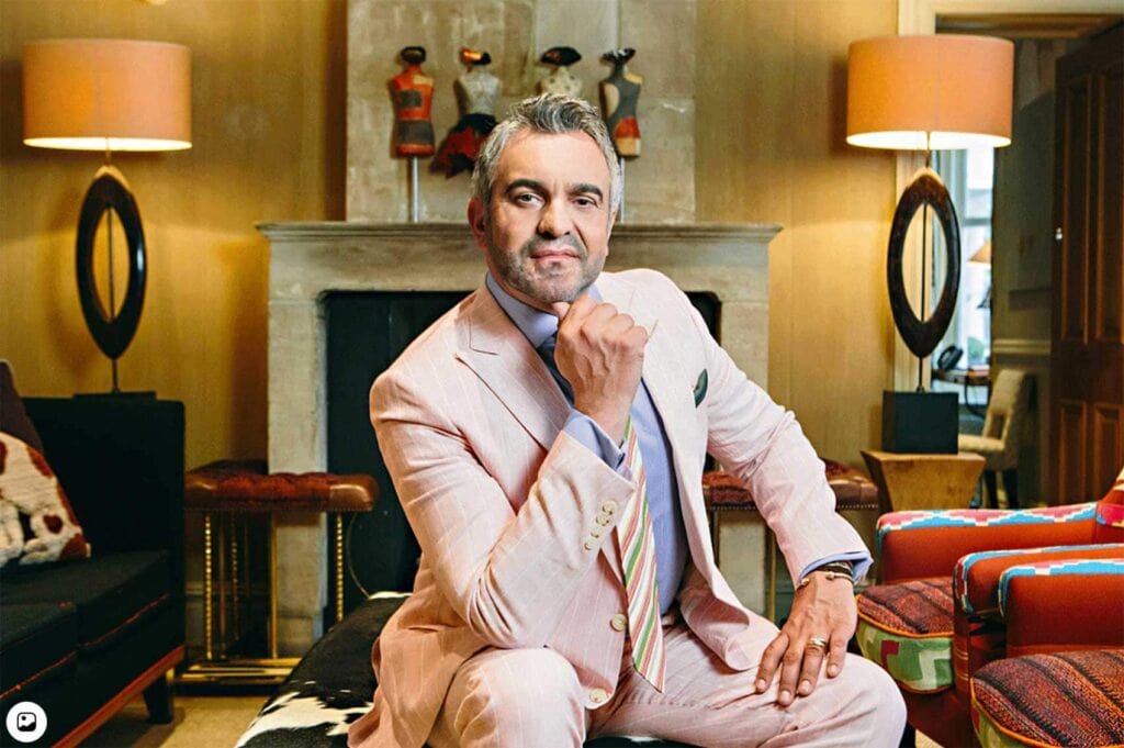 A man, Martyn Lawrence Bullard, in a light-pink suit poses seated with his fist beneath his chin in a room with lamps and a fireplace.