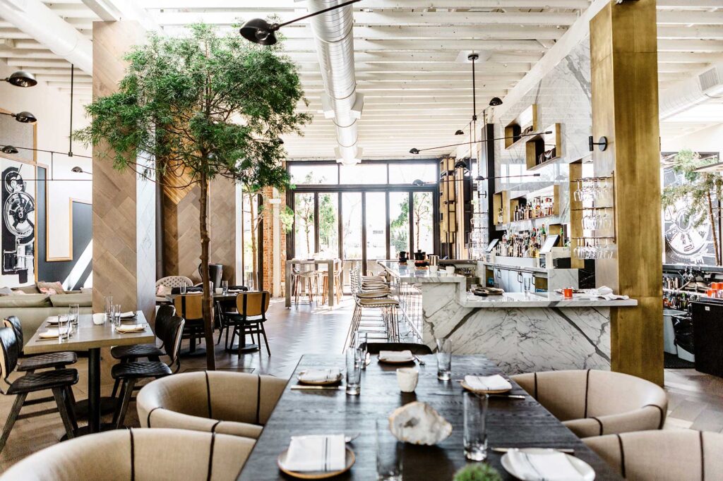 An airy, modern interior of a restaurant, with sleek black light fixtures, a marble bar, exposed ductwork and an indoor tree. Norah, West Hollywood, California.