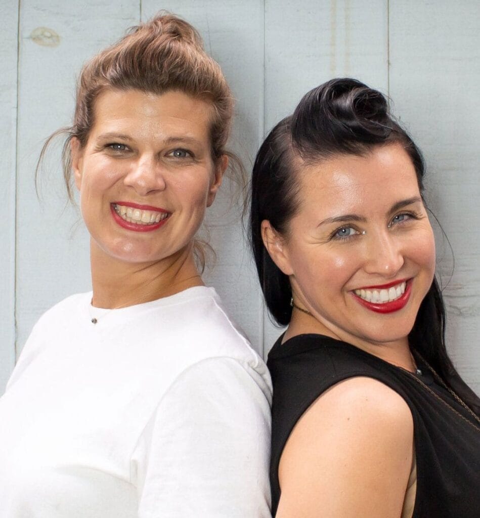 Two smiling women, Sarah Mengoni and Katie Brightside, stand back to back against a light blue wall.