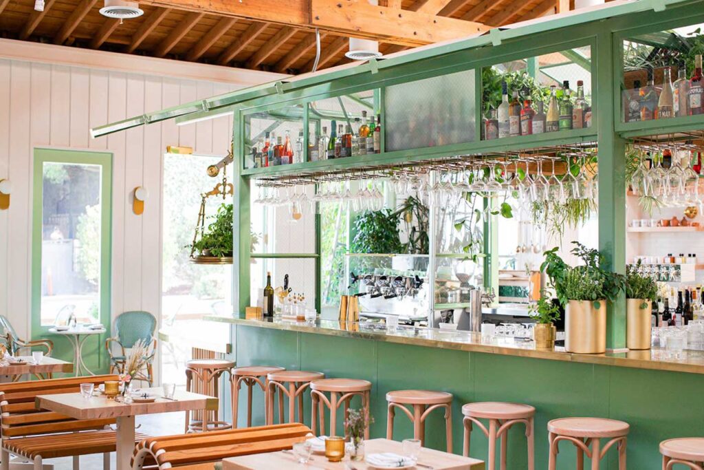 A light-filled restaurant featuring a green bar with wooden seats, white shiplap walls and gold fixtures. The Butcher's Daughter, West Hollywood, California.