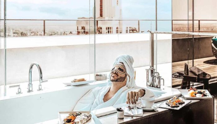 A man wearing a head wrap and cucumber slices on his eyes relaxes in a bathtub at a West Hollywood hotel.