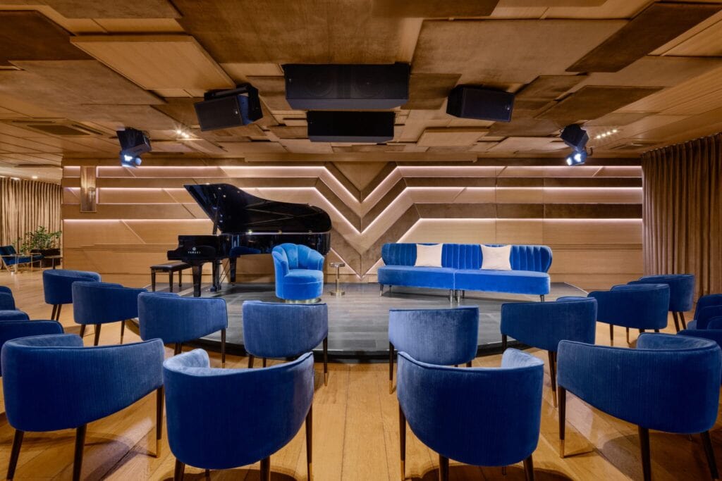 A small music venue with wooden floors and ceilings, blue velvet chairs and a grand piano on a small stage at The Sun Rose Hotel in West Hollywood, CA.