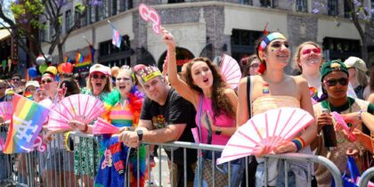 A diverse group of people stand behind a metal barricade at a pride parade, holding fans and rainbow flags, wearing colorful outfits and face paint, and cheering.