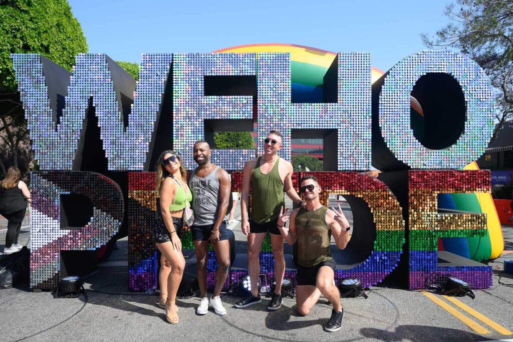Four people pose in front of a large, glittery "WEHO PRIDE" sign at the WeHo Pride event in CA.