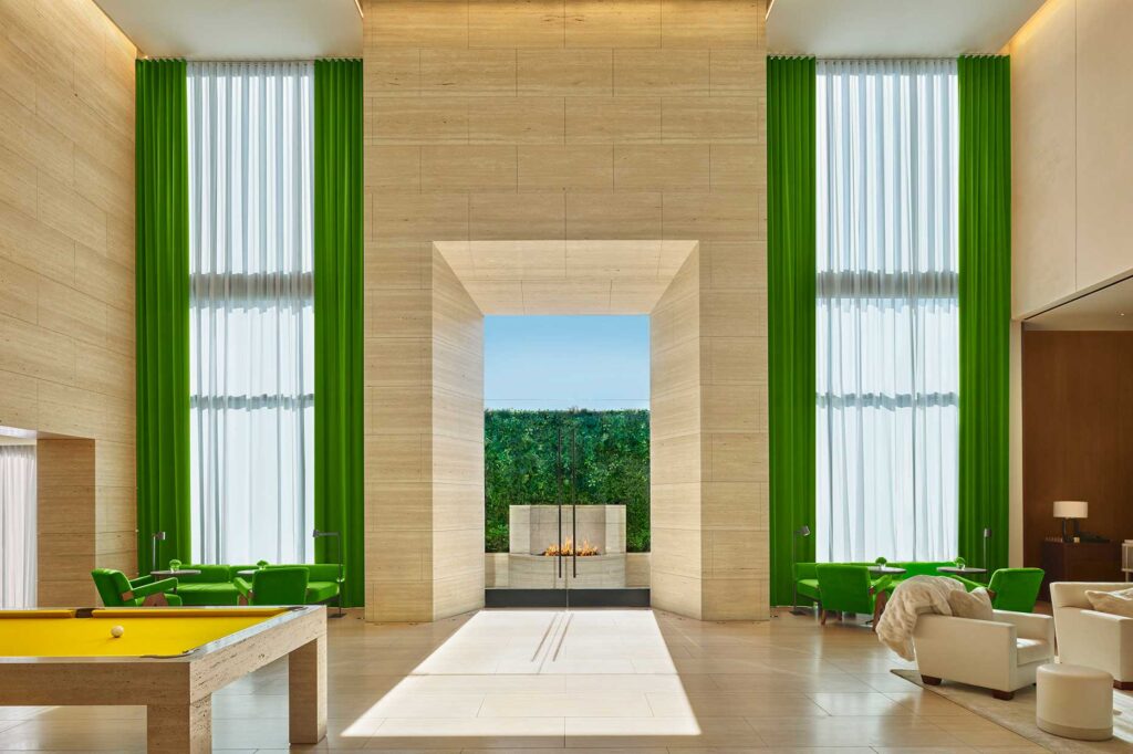 The lobby of the West Hollywood EDITION, with floor-to-ceiling windows, covered by bright green drapes, and large glass doors leading to an inviting outdoor space with a fireplace.
