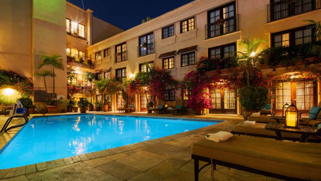 Night view of a hotel courtyard with illuminated swimming pool, surrounded by lounge chairs and vibrant plants.