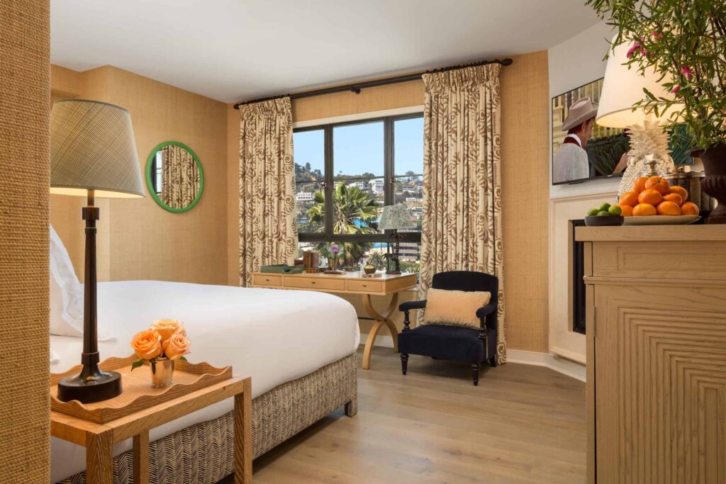 Well-lit hotel room with a neatly made bed, small desk by a window, cushioned chair and side table with flowers and fruit, featuring neutral tones and patterned curtains.