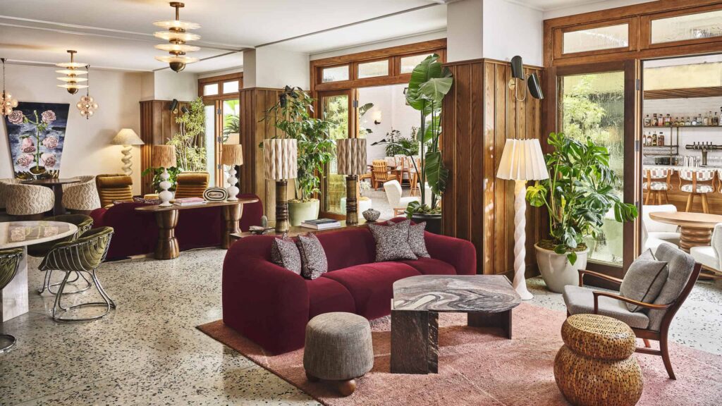 A stylish sitting room at Soho House Holloway in West Hollywood, CA, with a red curved sofa, decorative pillows, wooden coffee table, various lamps, indoor plants and window-doors letting in natural light.