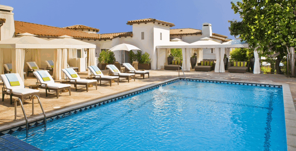 Outdoor swimming pool with blue water, surrounded by white lounge chairs, umbrellas and cabanas, set against a large, Mediterranean-style building on a sunny day.
