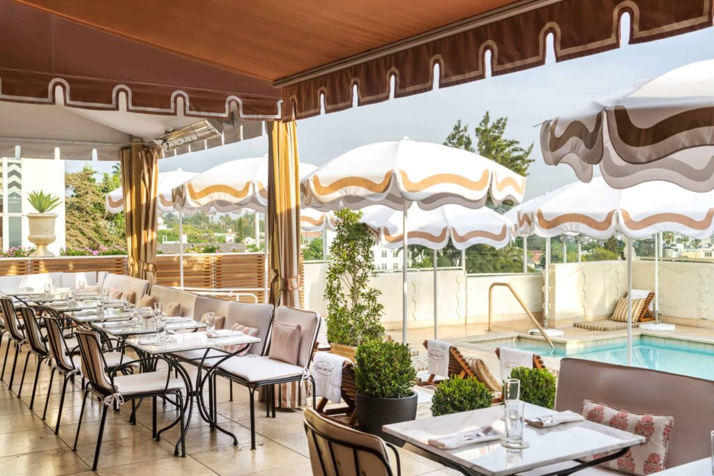 Outdoor dining area in West Hollywood with neatly set tables and chairs next to a pool, shaded by white umbrellas with brown trim on a sunny day.