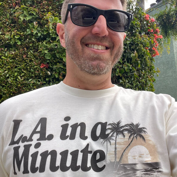Close-up selfie of a man wearing black sunglasses and wearing a shirt that reads "L.A. in a Minute." West Hollywood, California.