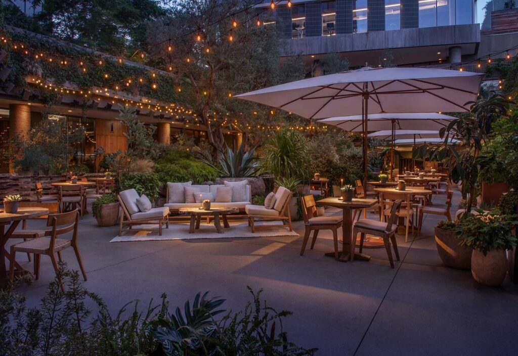 A lush courtyard at the 1 Hotel West Hollywood pictured at dusk features neutral-colored couches, dining tables with umbrellas, a variety of foliage and string lights. West Hollywood, California.