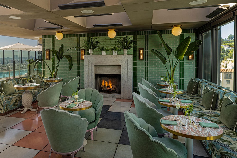 Upscale interior of The Rooftop Bar at The Sun Rose in West Hollywood with green tiled walls, fireplace, green chairs, round tables, potted plants, and large windows offering views of a cityscape.