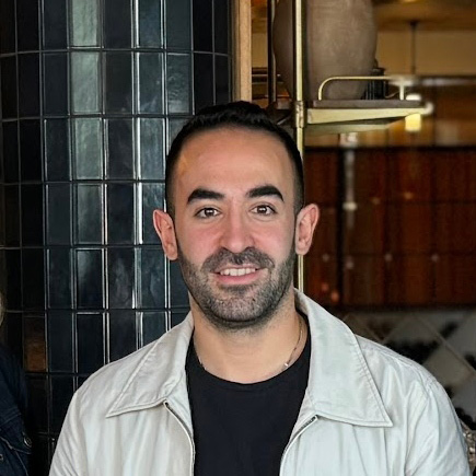 A man wearing a black t-shirt and white jacket smiles at the camera. Charbel Hayek of Ladyhawk, West Hollywood, California.