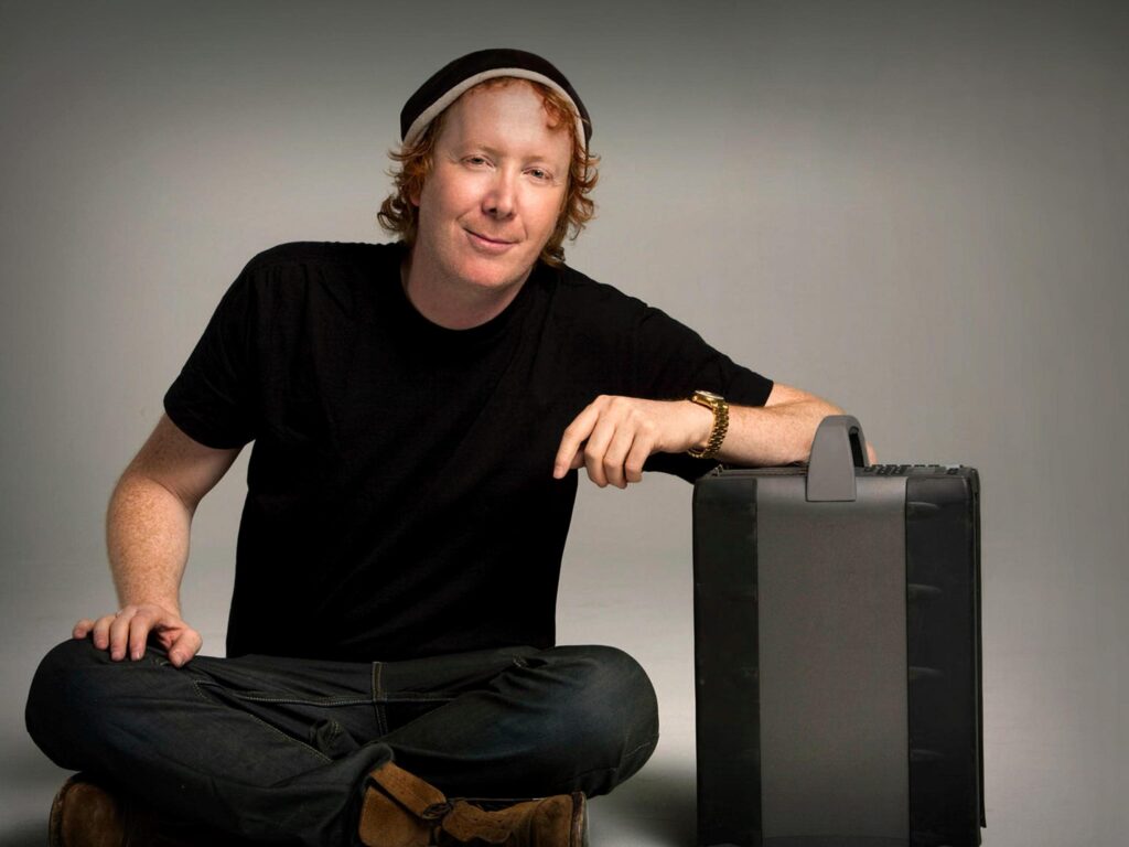 Celebrity photographer Scott Nathan, wearing all black clothing and a beanie hat, sits cross-legged in a portrait.