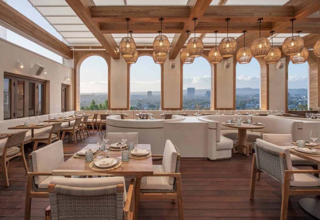 Arched windows at Casa Madera in West Hollywood, CA, reveal Los Angeles views beside white dining chairs, wooden tables and chandeliers overhead.